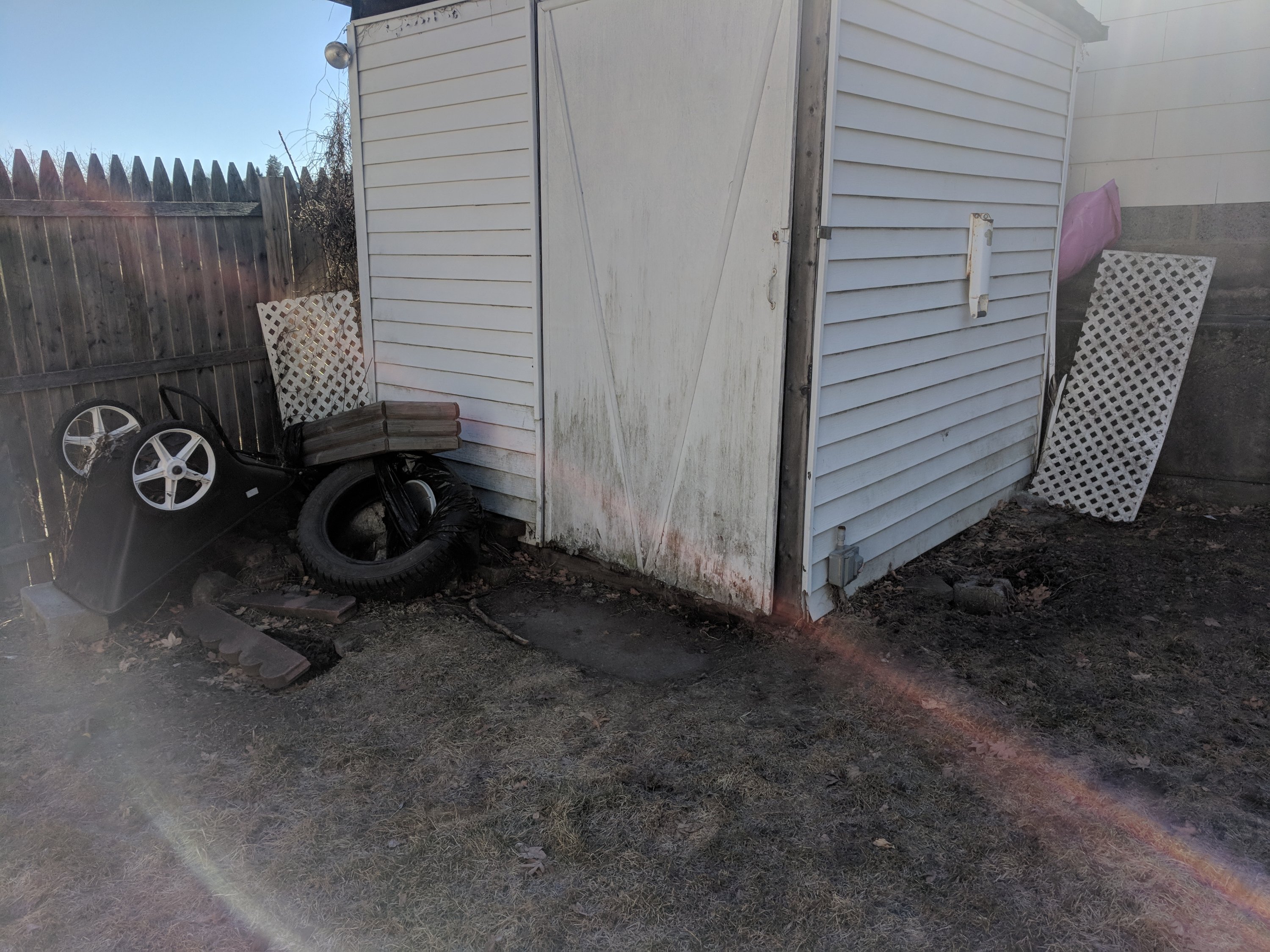 A small white shed with a fenced yard. Tires, a tilted cart, and lattice panels are leaning against the shed.