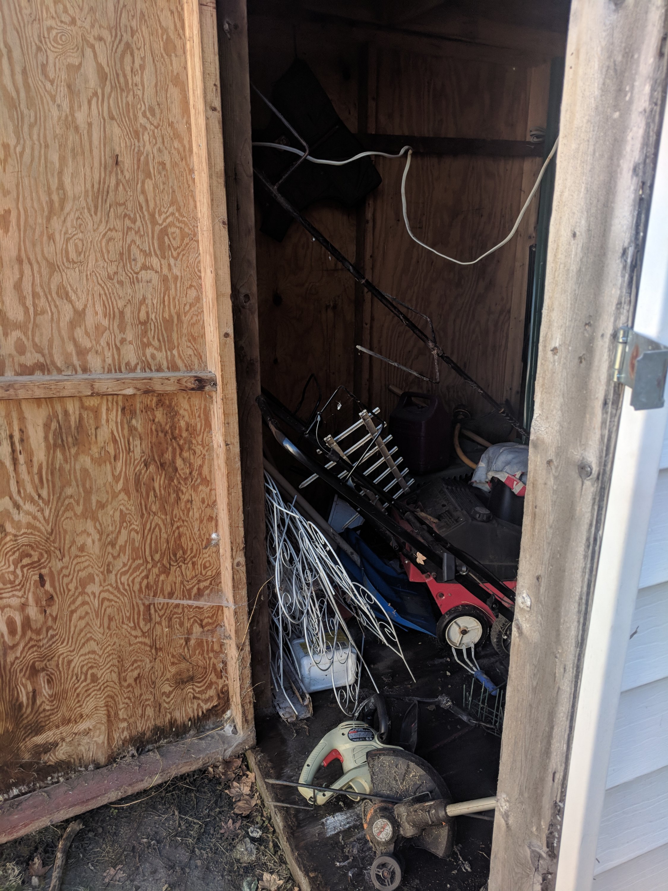 A cluttered shed with garden tools, a lawn mower, coiled wires, and various yard equipment.