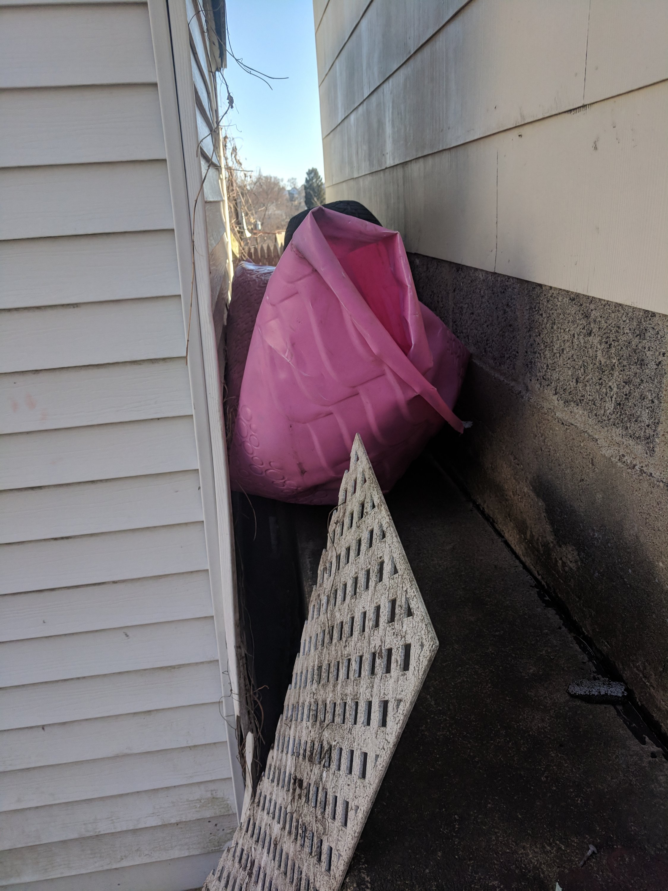 A large pink inflatable object wedged between two building walls. A white lattice panel lies on the ground in the foreground.