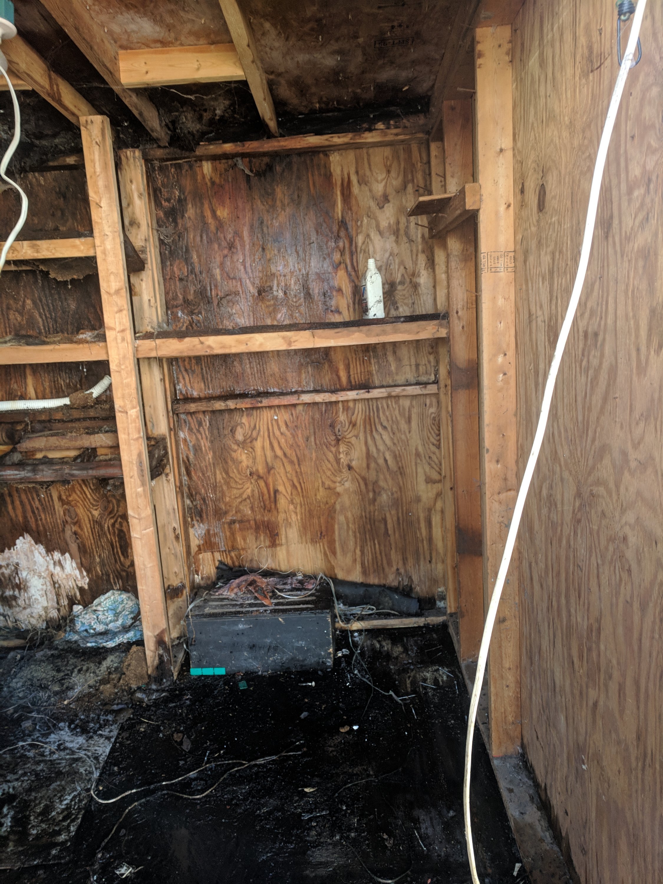 A charred wooden room with damaged walls and ceiling, exposed wires, and debris on the floor. Burn marks are visible throughout the space.