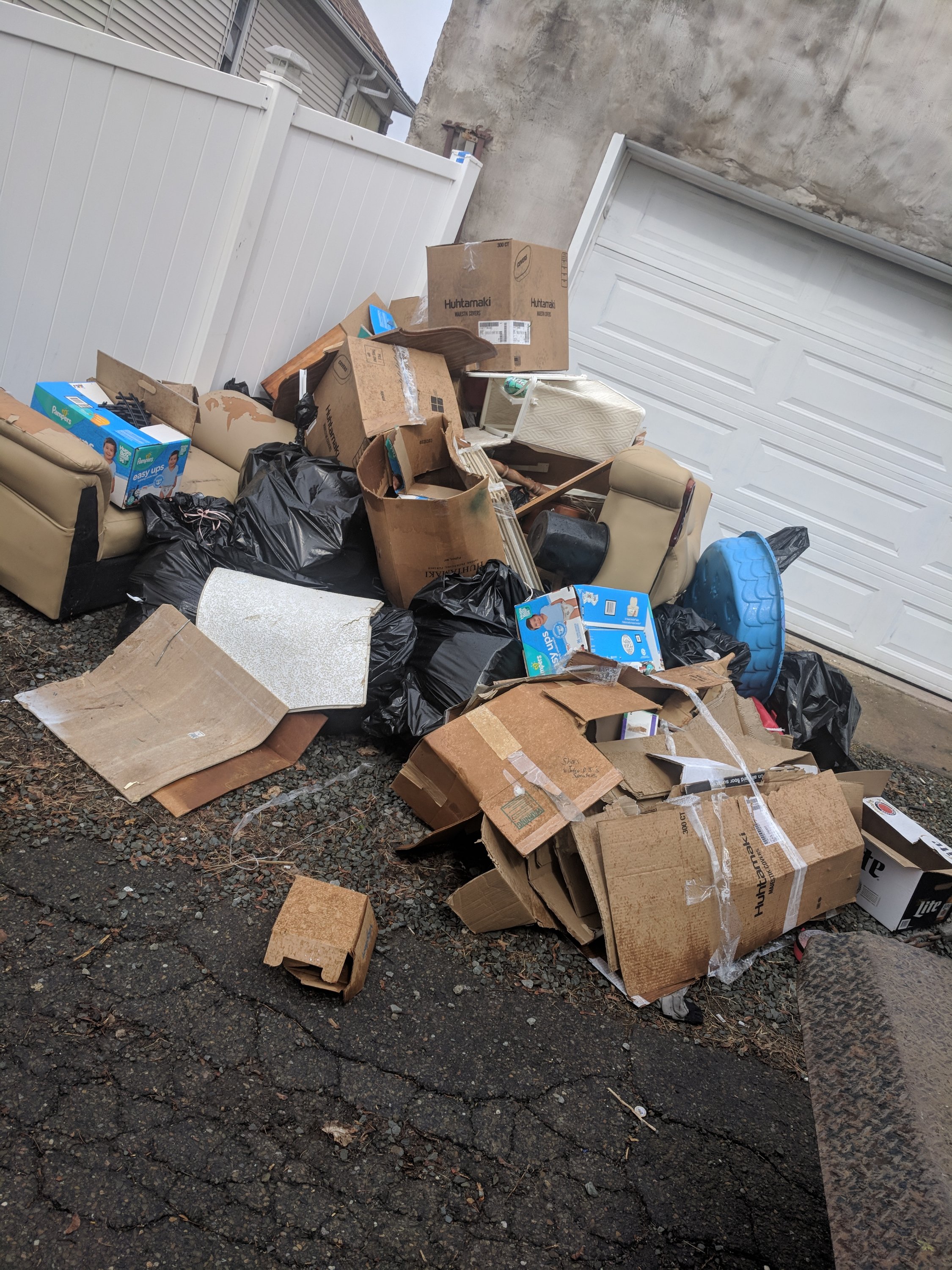 A large pile of cardboard boxes, black garbage bags, and miscellaneous discarded items is stacked near a white garage door.