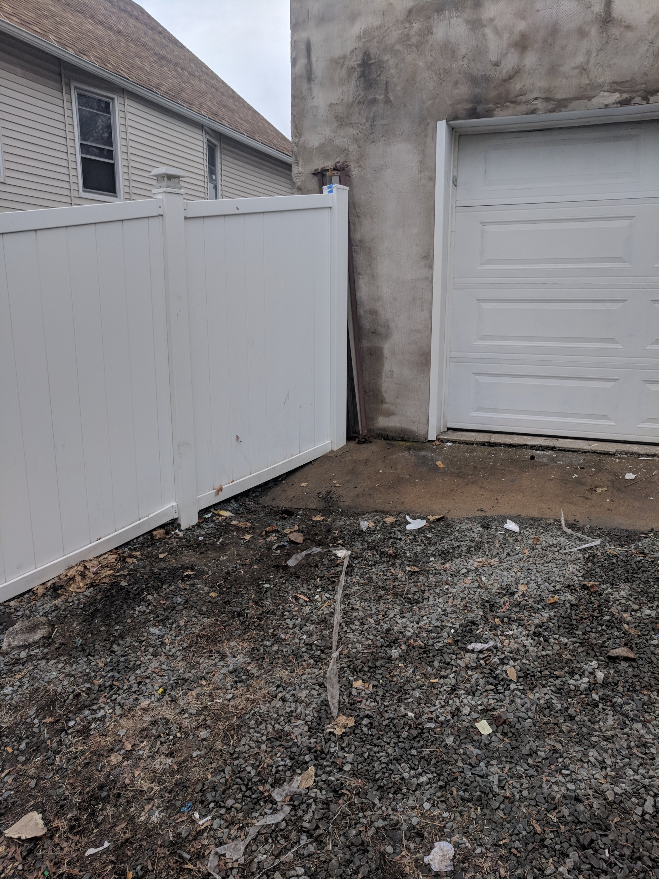 White fence gate partially open next to a white garage door. Ground is covered with gravel and debris, and two house exteriors are visible.