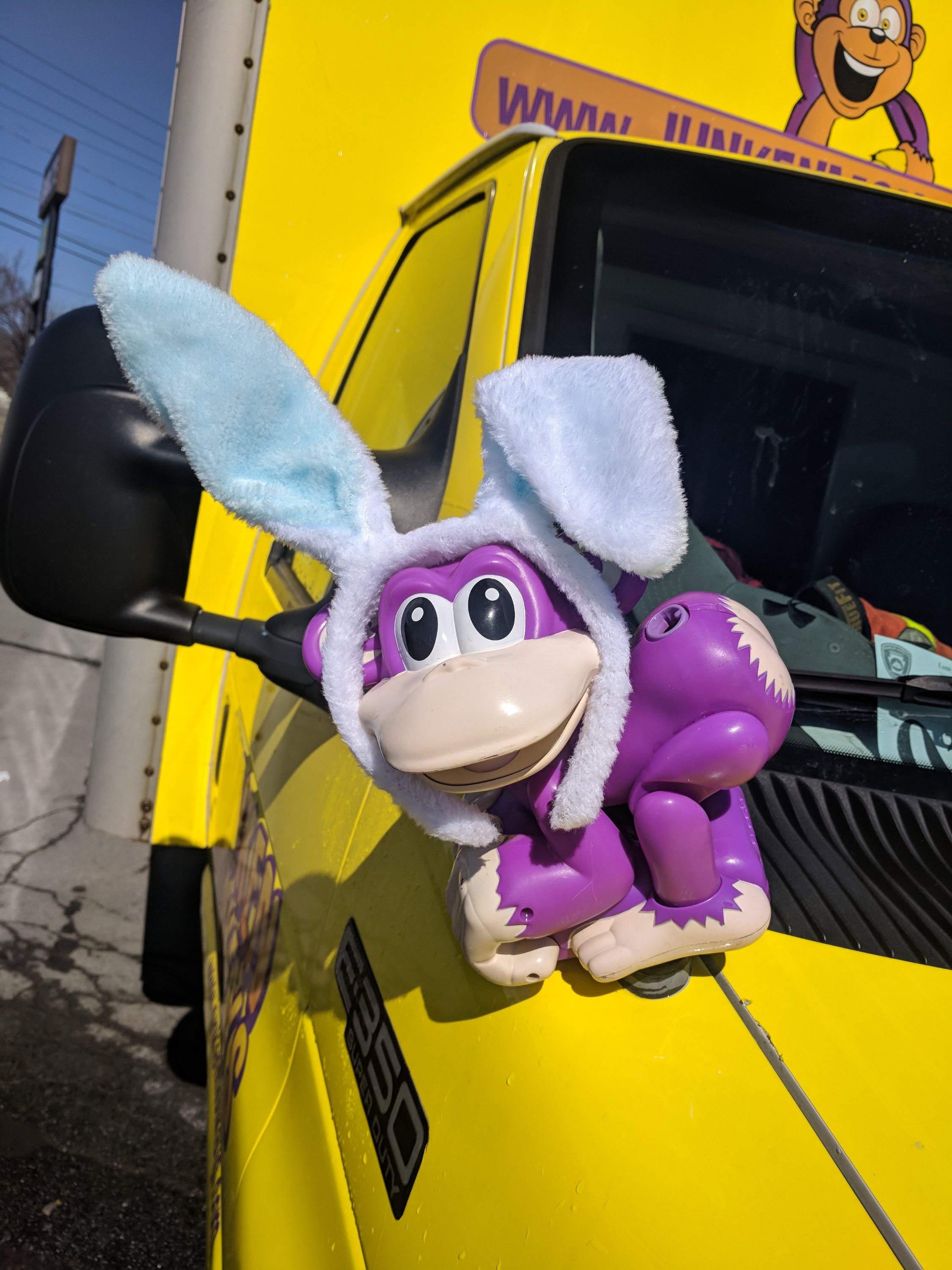 Purple monkey toy with blue and white bunny ears, attached to the side mirror of a yellow truck featuring cartoon monkey graphics.