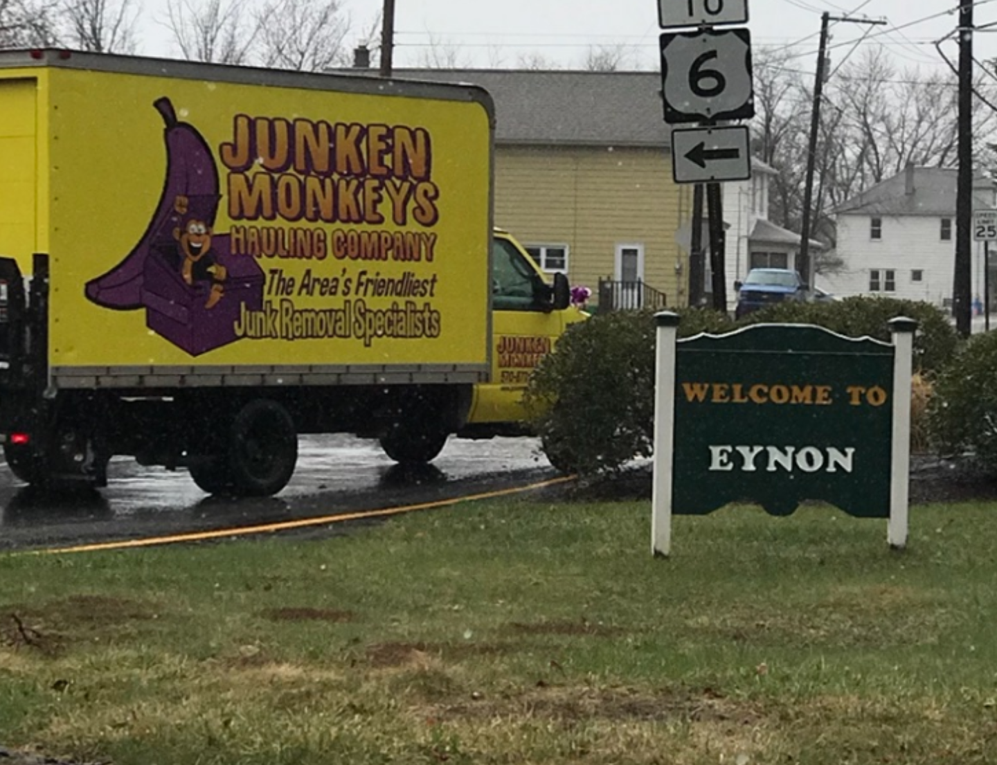 A yellow truck with "Junken Monkeys Hauling Company" is near a sign reading "Welcome to Eynon" beside a road on a rainy day.