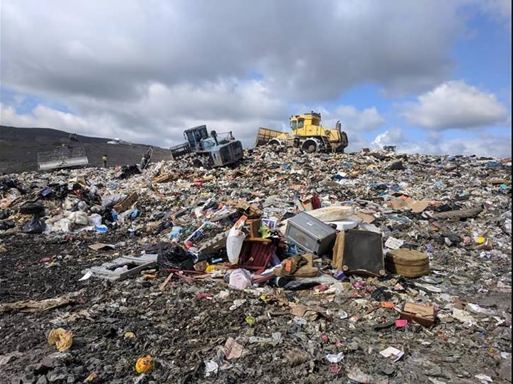 A landfill with piles of garbage, including broken furniture and household items, under a cloudy sky. Two vehicles are on top, possibly moving debris.