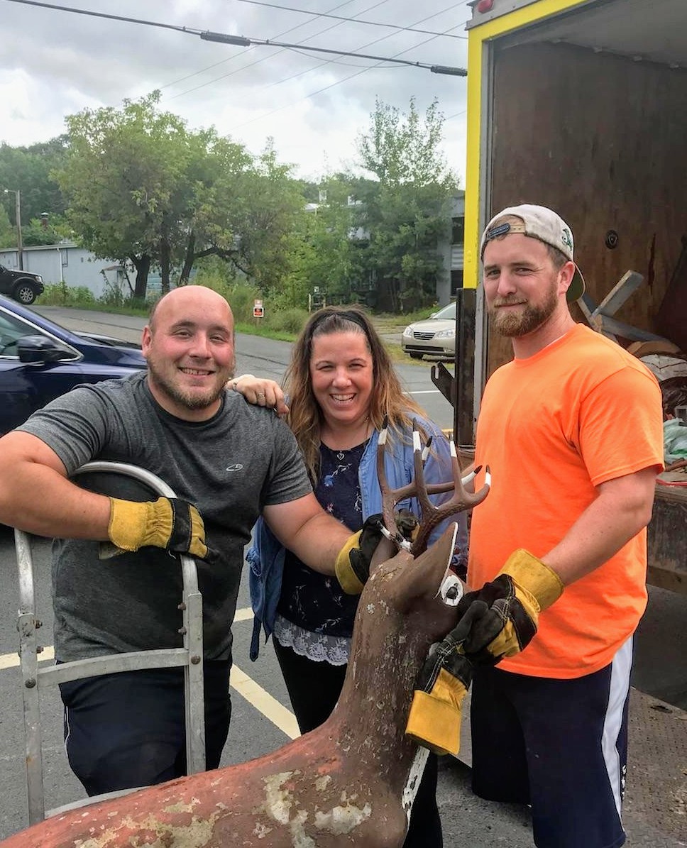 Three people posing with a deer statue near a moving truck; two men in work gloves, and one woman smiling in the middle.