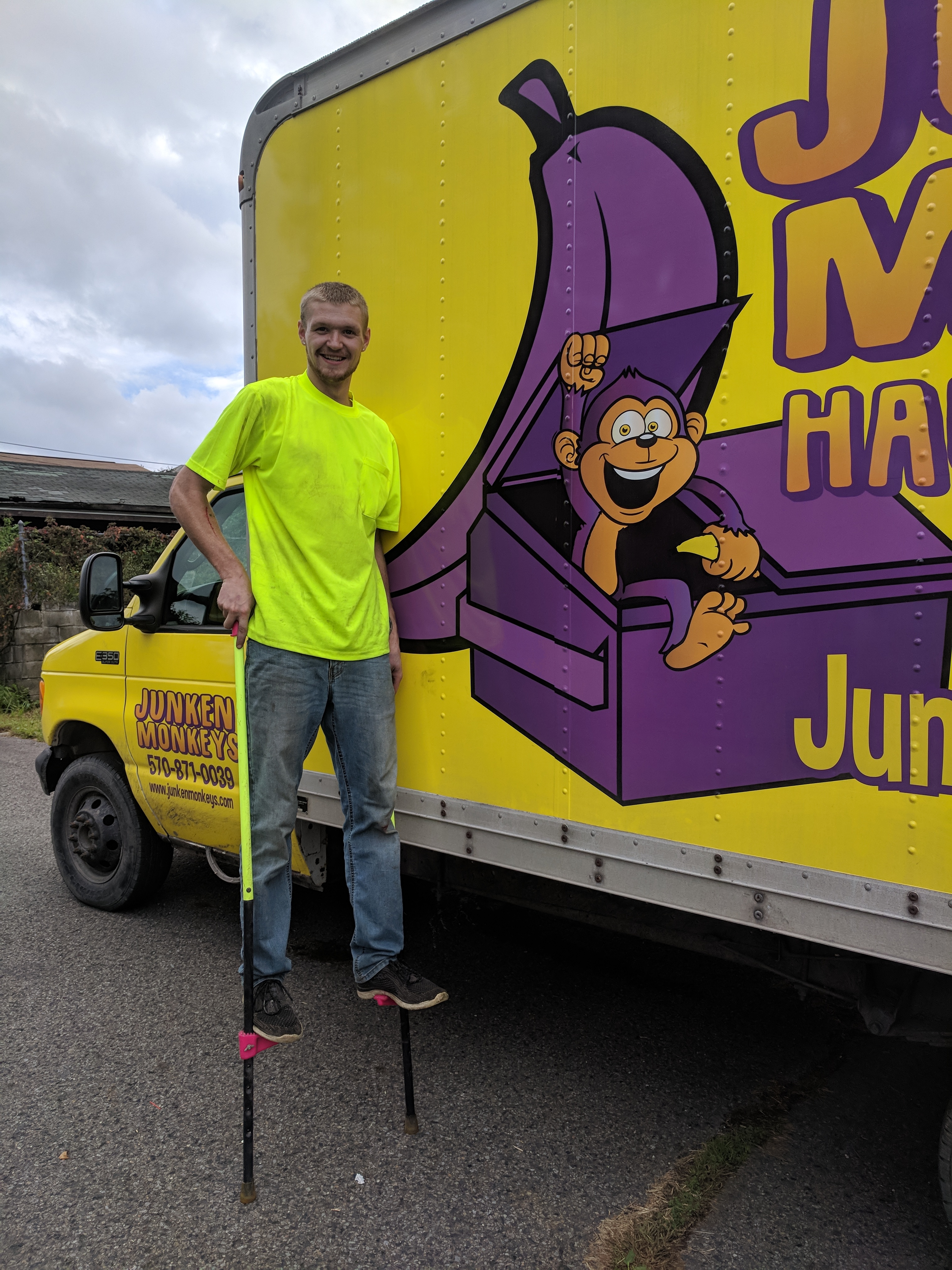 A man in a neon yellow shirt stands on stilts next to a colorful truck with a cartoon monkey on the side.