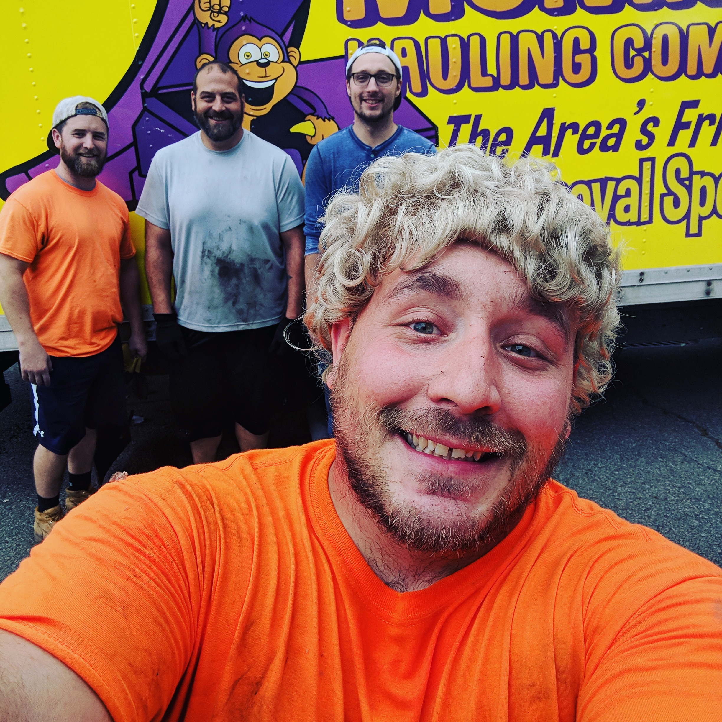 A group of four men pose in front of a yellow and purple truck, with one man taking a selfie. They are wearing casual work clothes.