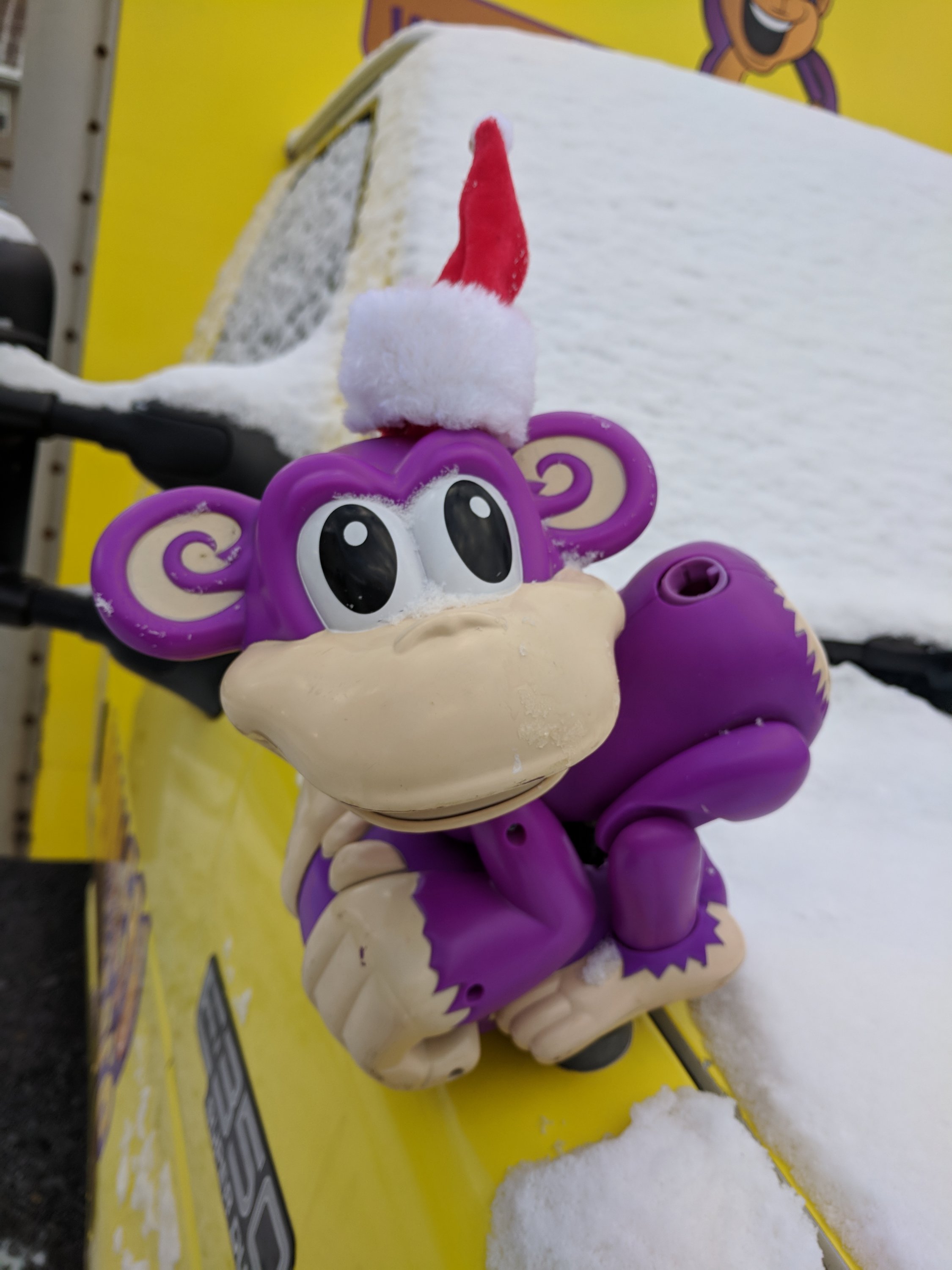 Purple toy monkey with large eyes and a Santa hat is mounted on a snow-covered yellow vehicle.