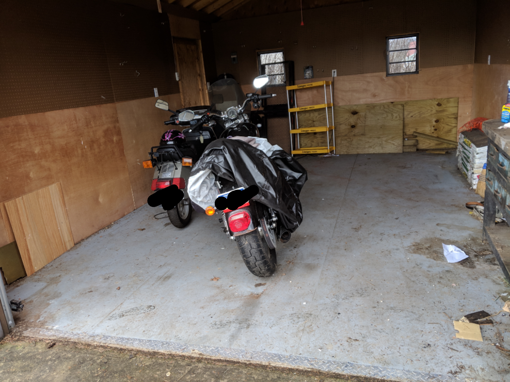 Two motorcycles parked inside an empty garage with a ladder and tools against the wall.