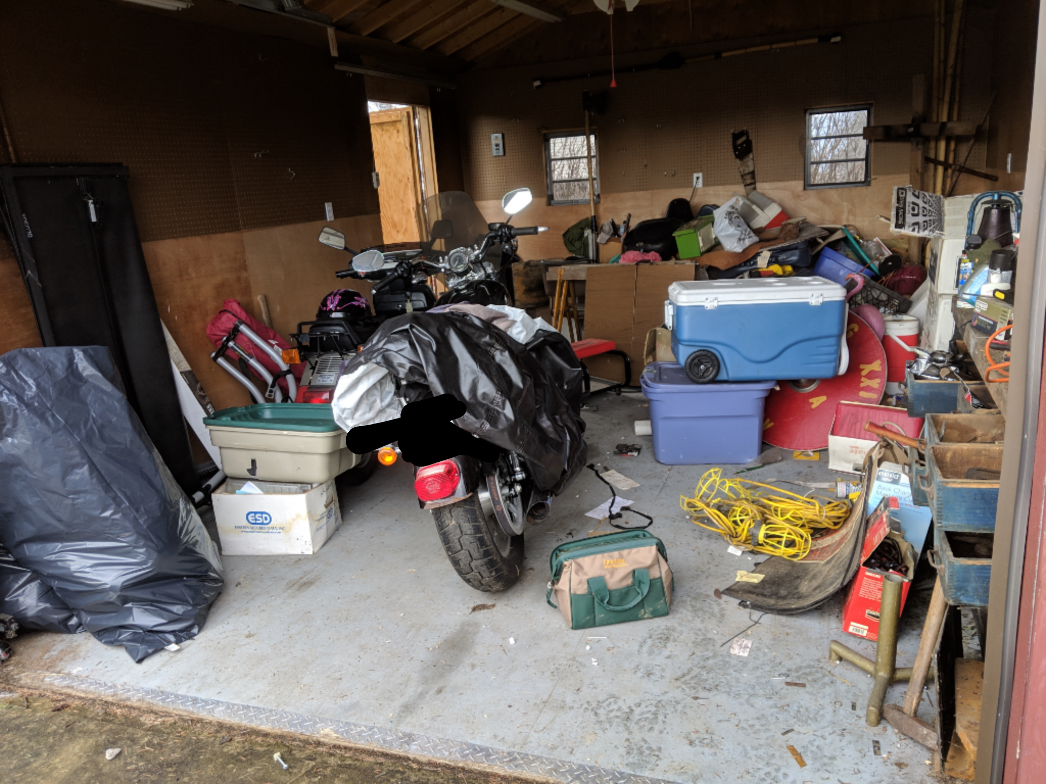 A cluttered garage with a motorcycle covered by a tarp, surrounded by various items including coolers, bags, and tools.