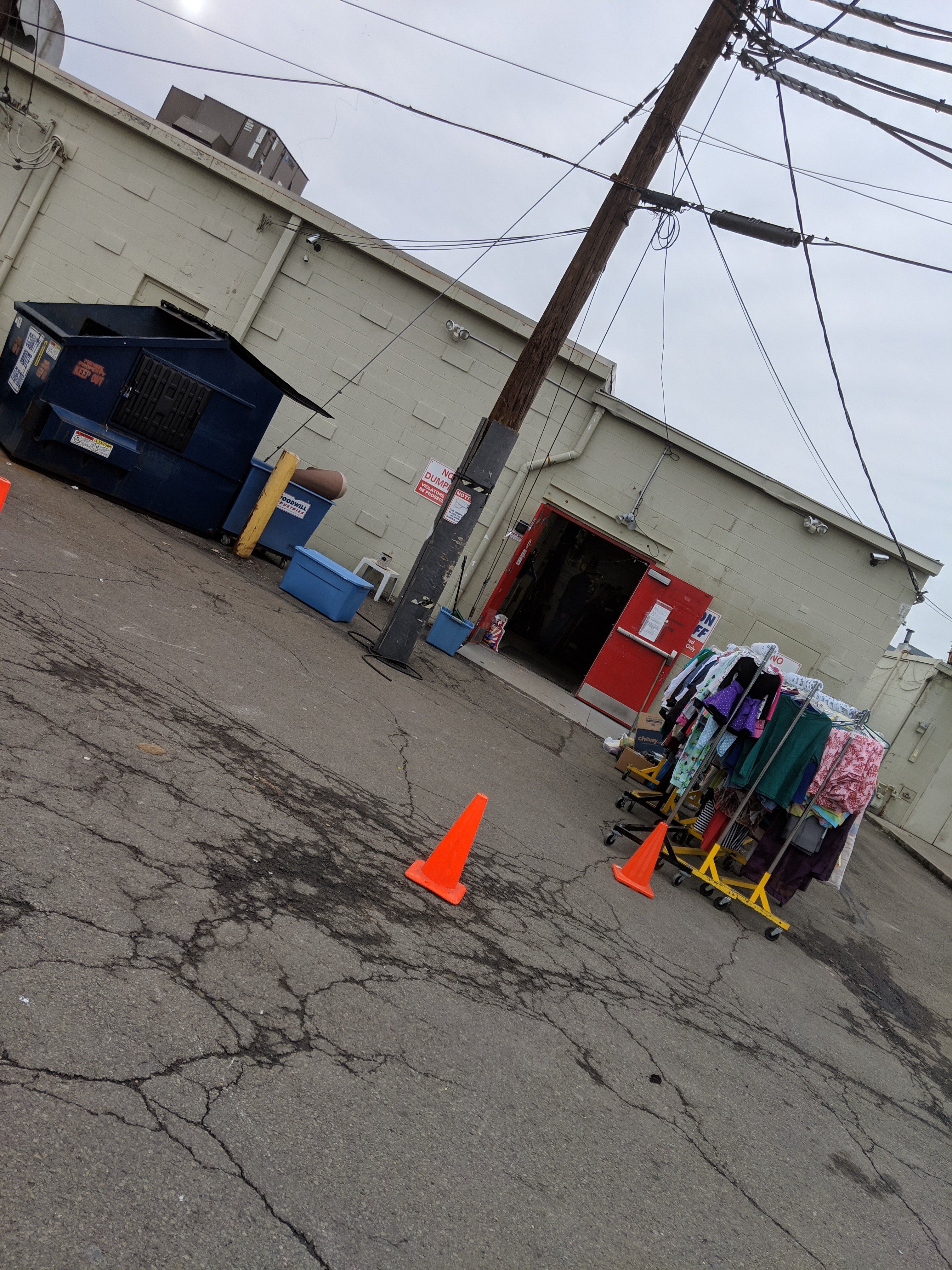 Clothes displayed on racks outside a building near a dumpster. Orange cones are placed on the cracked pavement. Overhead power lines are visible.