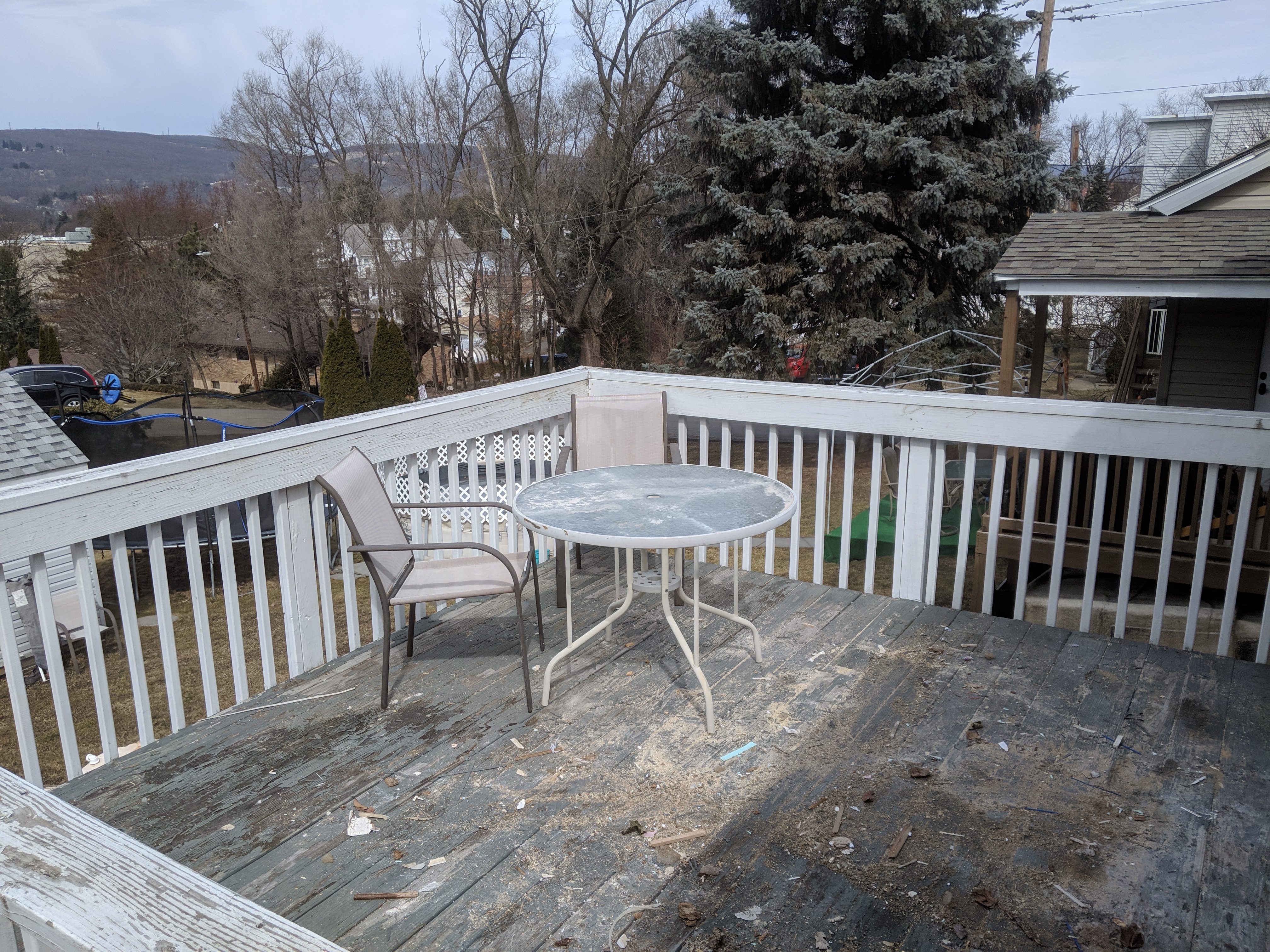 Weathered wooden deck with a round metal table and two chairs, surrounded by a white railing. Trees and neighboring houses are visible in the background.