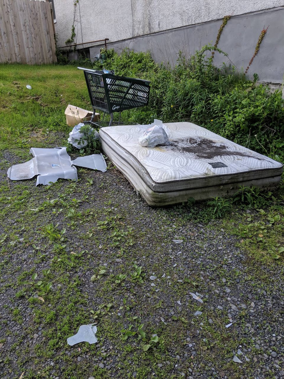 Discarded mattress, broken plastic pieces, and a shopping cart on a gravel and grass lot near a building.