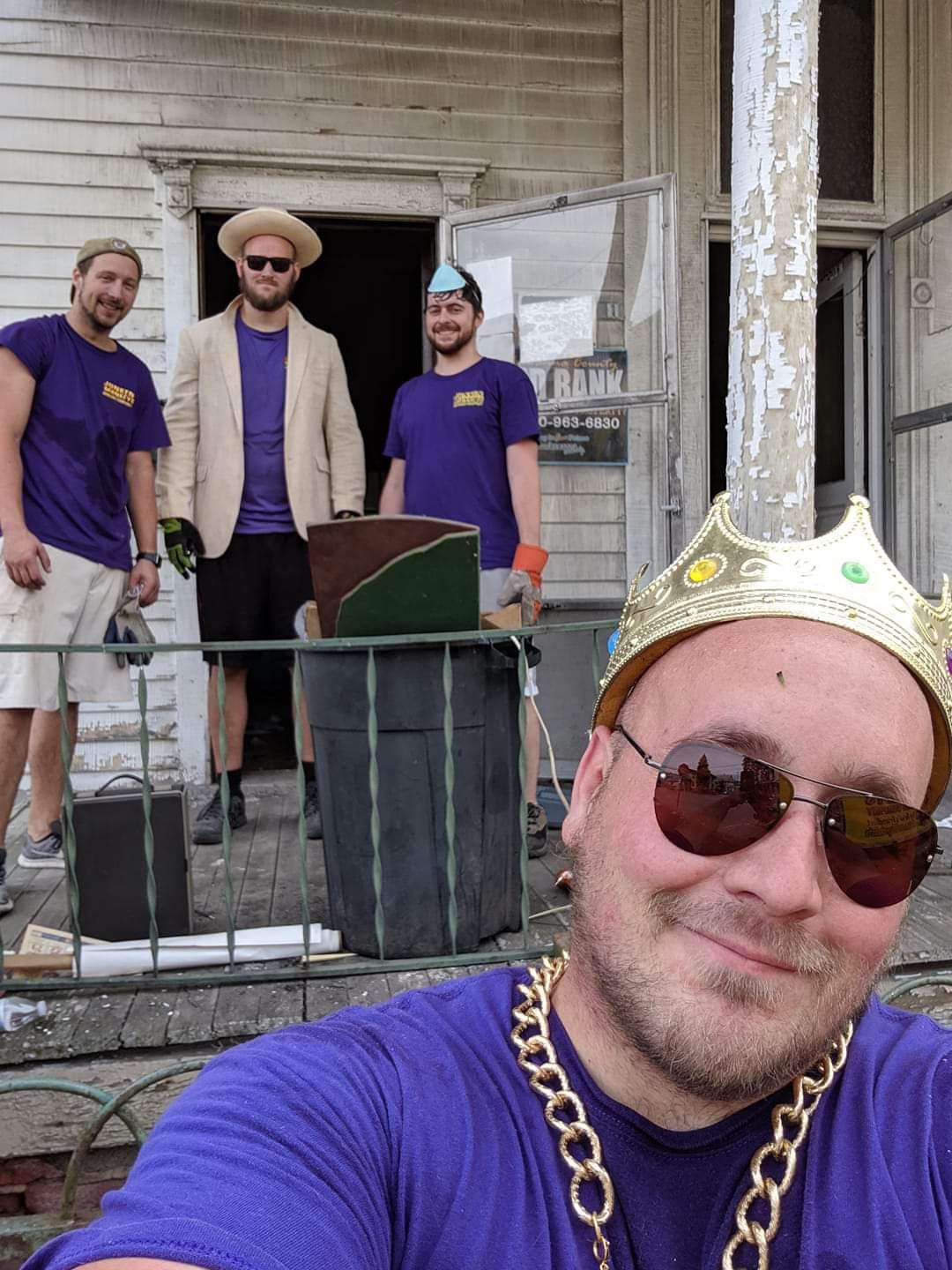 Four people wearing purple shirts stand and sit on a porch. One person in the foreground takes a selfie, wearing a crown and chain necklace. The house shows signs of wear.