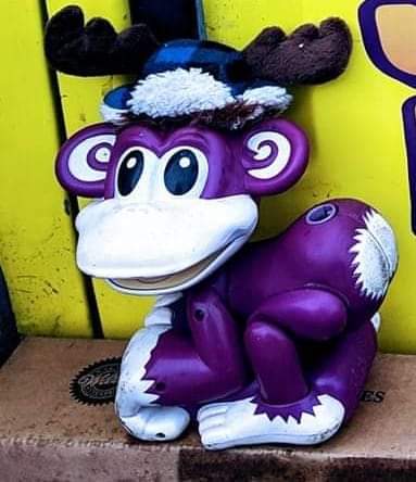 A purple monkey toy with antlers and a blue hat sits on a cardboard box against a yellow background.