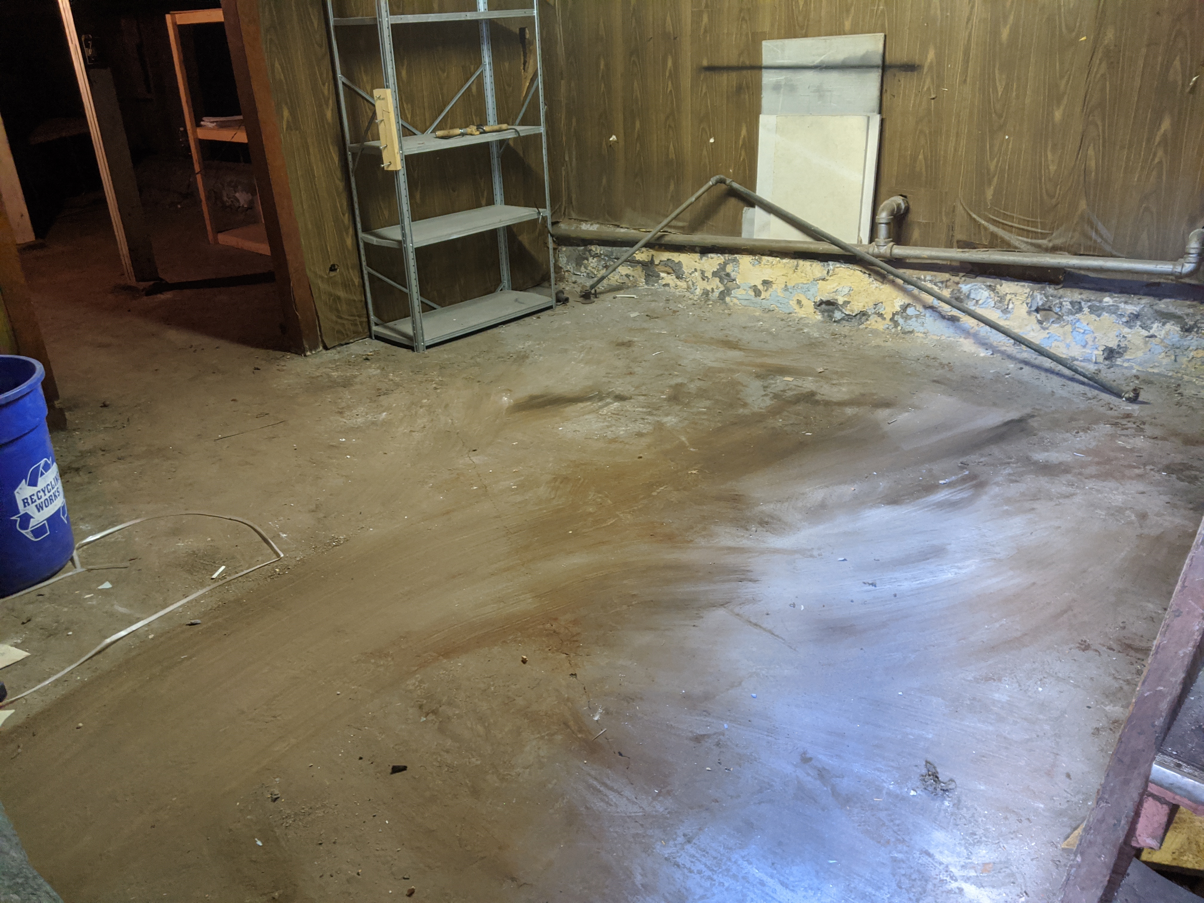 Empty basement corner with bare concrete floor, a metal shelf, a blue bucket, and exposed pipes on wood-paneled walls.