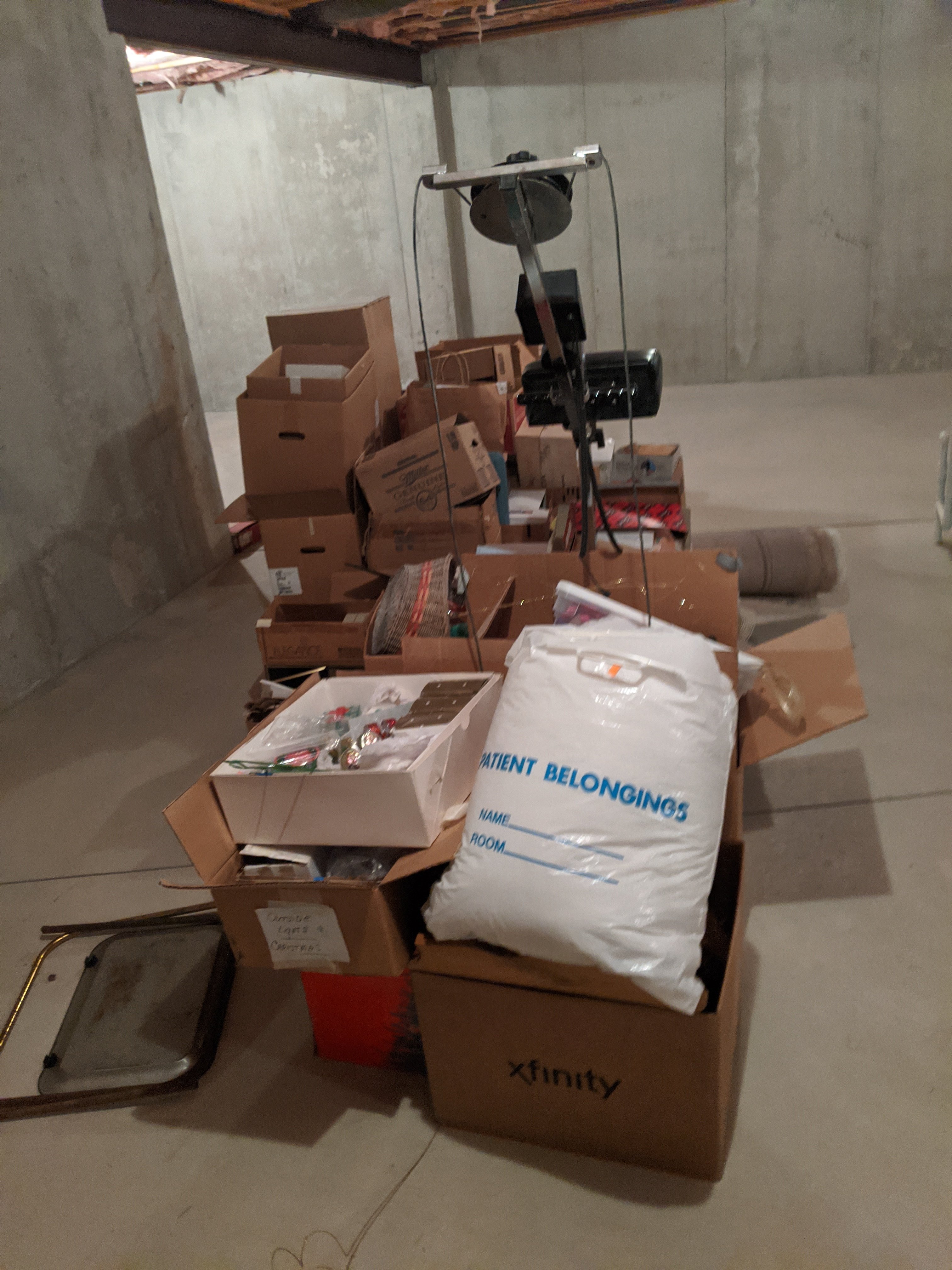 A cluttered basement with cardboard boxes, a vacuum cleaner, and a large "Patient Belongings" bag on a concrete floor.