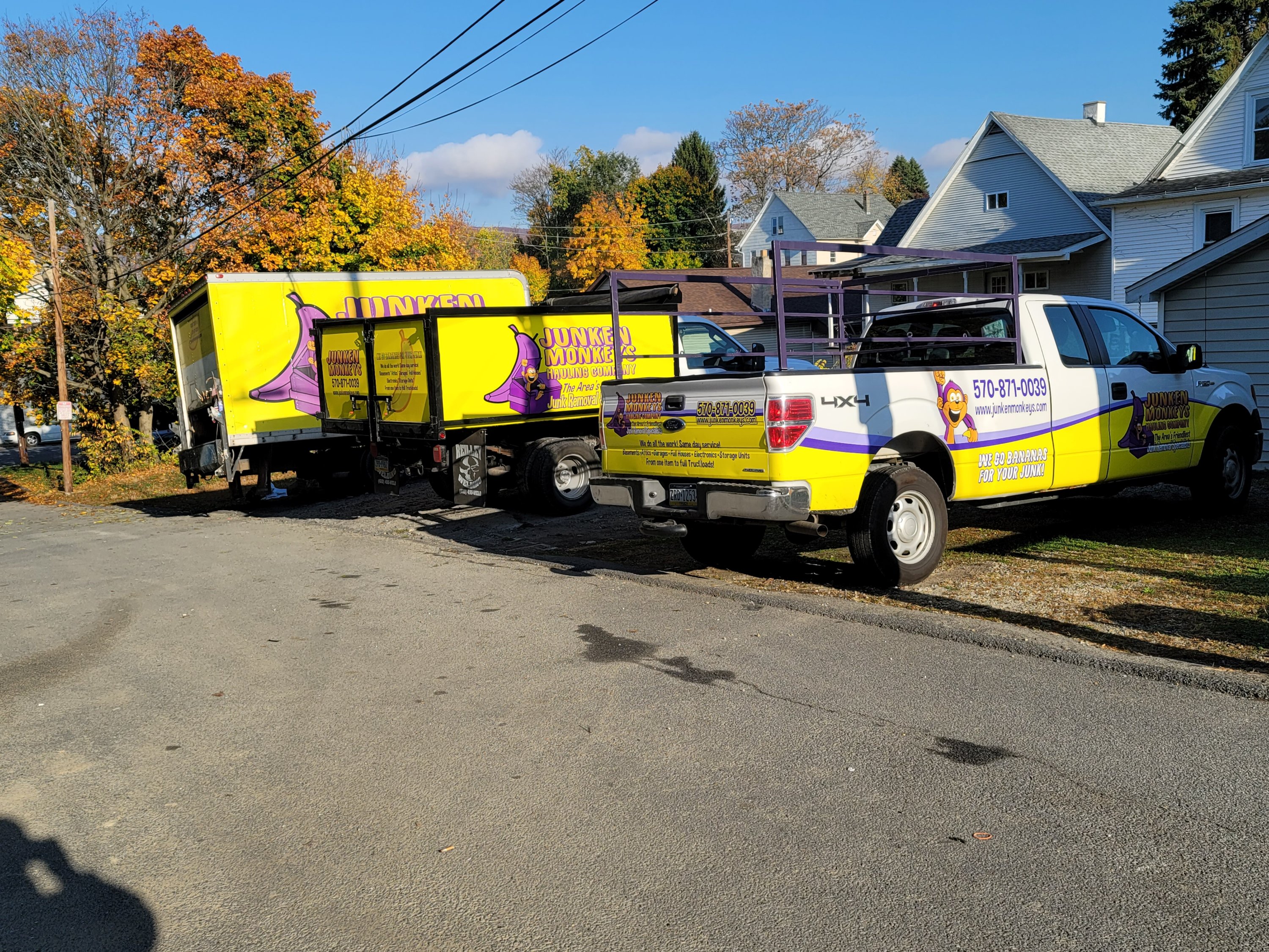 Two yellow and purple trucks labeled "Junk King" parked on a residential street, surrounded by autumn trees and houses.