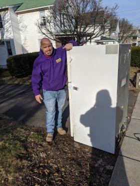 A man in a purple hoodie stands on a sidewalk, leaning on a large white appliance, possibly a fridge, with a residential house in the background. A shadow of the photographer is visible.